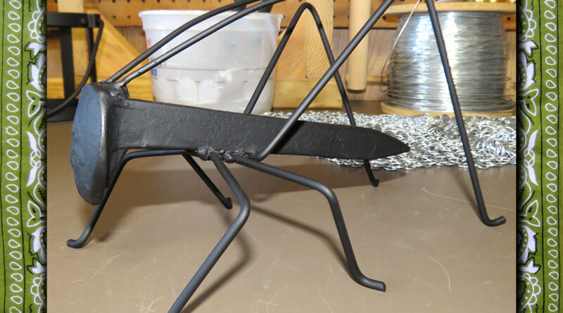 A welded steel cricket or grasshopper made from a railroad spike and steel rod; painted matte black; sitting on a work table.